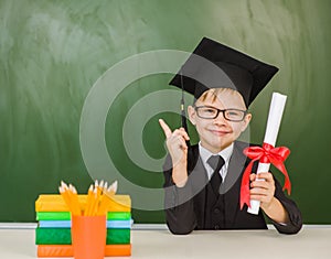 Happy boy with diploma in graduation hat points on empty green chalkboard