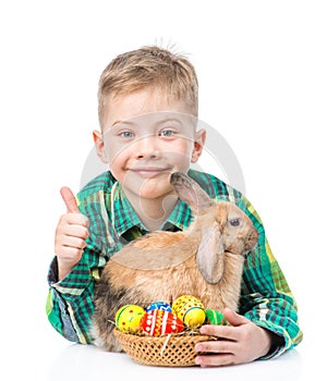 Happy boy with easter eggs embracing rabbit and showing thumbs up