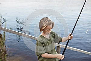 Happy boy fishing from wooden placing, holding fishing rod in hands and being ready to cast line, blonde guy spending time near