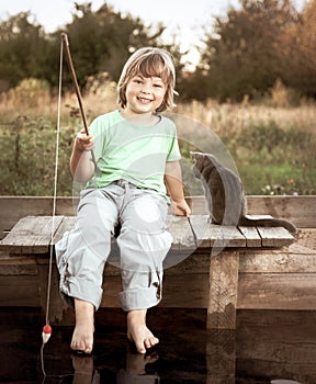 Happy boy go fishing on the river with pet, one children and kitten of the fisherman with a fishing rod on the shore of the lake.