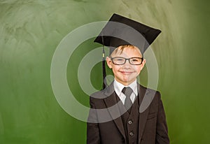 Happy boy in graduation cap standing near green chalkboard