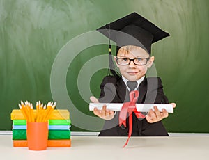 Happy boy in graduation hat gives a diploma near empty green chalkboard