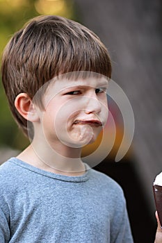 Happy boy in grey t shirt making faces and twisting lips while eating ice cream outdoor in the park. Close up funny face