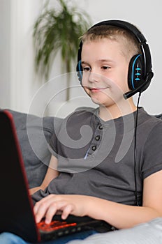 Happy boy in headphones playing online video game on laptop computer at home. Child sitting and use pc