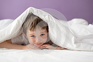 Happy boy hiding in bed under a white blanket or coverlet photo