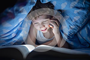 Happy boy is lying under blanket and reading book with flashlight, at night