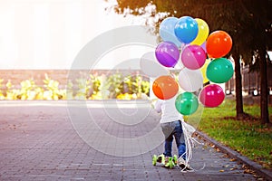 Happy boy playing with a bunch of balloons outside and riding a scooter. Back view