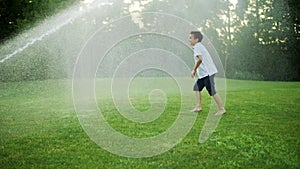 Happy boy playing with water sprinkler in field. Positive guy jumping in air