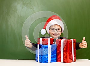 Happy boy in red christmas hat with gift boxes near empty green blackboard showing thumbs up