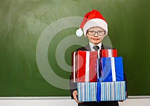 Happy boy in red christmas hat with gift boxes standing near empty green blackboard