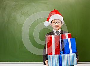 Happy boy in red christmas hat with gift boxes standing near empty green blackboard