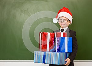 Happy boy in red christmas hat with gift boxes standing near empty green blackboard