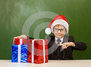 Happy boy in red christmas hat near empty green chalkboard points on on the gift boxes
