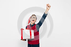 Happy boy in a red Santa hat. Stylish boy with Christmas gift having fun and showing gesture of victory and success on white