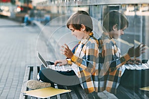 A happy boy is sitting on a bench, holding a laptop on his lap. The boy communicates with friends using the computer, waving to