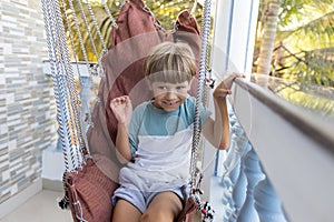 Happy boy is sitting, swinging in rattan swing chair during summer holidays at hotel having fun.