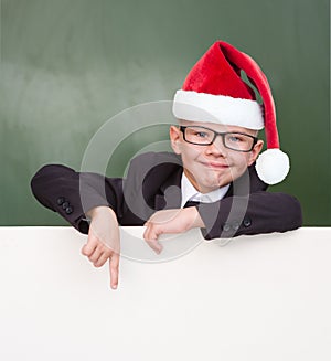 Happy boy in a suit with red christmas hat pointing down at blank billboard