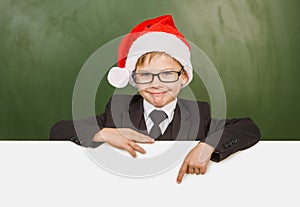 Happy boy in a suit with red christmas hat pointing down at blank billboard