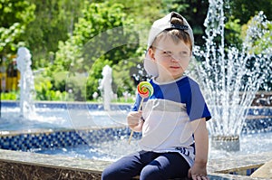 happy boy of two years is sitting near the fountain in the summer and eats a delicious multi-colored candy on a stick.