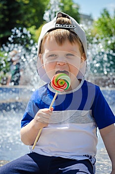 happy boy of two years is sitting near the fountain in the summer and eats a delicious multi-colored candy on a stick.