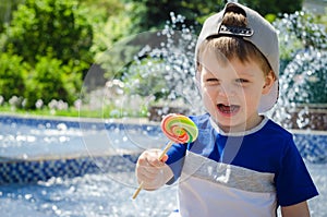 happy boy of two years is sitting near the fountain in the summer and eats a delicious multi-colored candy on a stick.