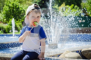 happy boy of two years is sitting near the fountain in the summer and eats a delicious multi-colored candy on a stick.