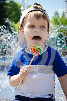 happy boy of two years is sitting near the fountain in the summer and eats a delicious multi-colored candy on a stick.