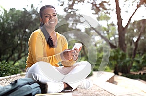 Happy brazilian college lady using cellphone, sitting in park or university campus, smiling at camera, copy space