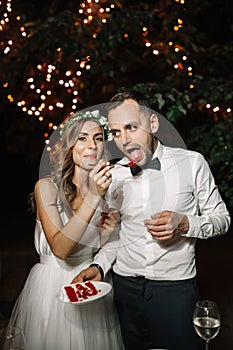 Happy bride and groom cut the wedding cake in the front garland light decoration. Wedding evening outdoor.