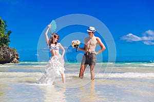 Happy bride and groom with a fan and a wedding bouquet having fun on the beach on the tropical island.