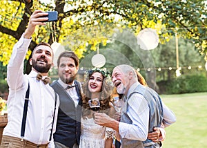 Bride, groom and guests with smartphone taking selfie outside at wedding reception.