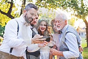 Bride, groom and guests with smartphones taking selfie outside at wedding reception.