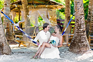 Happy bride and groom having fun in a hammock on a tropical beach under the palm trees. Summer vacation concept.