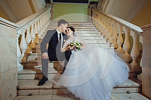 Happy bride and groom are sitting on a white marble staircase, holding hands and looking into each other`s eyes