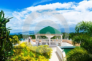 Happy bride and groom standing next to the stone gazebo amid beautiful tropical landscape. Sea, sky, flowering plants and palm tr