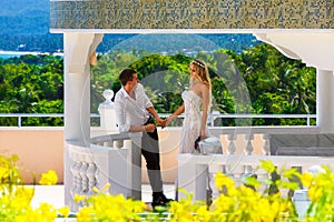Happy bride and groom standing next to the stone gazebo amid beautiful tropical landscape. Sea, sky, flowering plants and palm tr