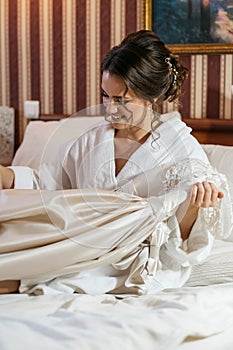 A happy bride is sitting on the bed and is holding her wedding dress