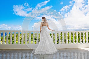 Happy bride standing next to the stone gazebo amid beautiful tropical landscape. Sea, sky, flowering plants and palm trees in