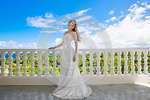 Happy bride standing next to the stone gazebo amid beautiful tropical landscape. Sea, sky, flowering plants and palm trees in