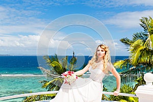 Happy bride standing next to the stone gazebo amid beautiful tropical landscape. Sea, sky, flowering plants and palm trees in