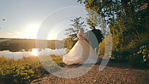 Happy bride in white dress, and groom walking down country road, holding hands in setting sun. Back view of wedding