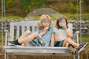 Happy brother and sister swinging on swing in summer park outdoors. Little boy and girl kids drink green smoothie