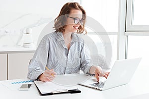 Happy brunette businesswoman in glasses using laptop computer while working in light apartment