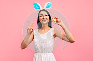 Happy brunette girl in bunny ears and with painted easter eggs showing two fingers gesture victory, easter