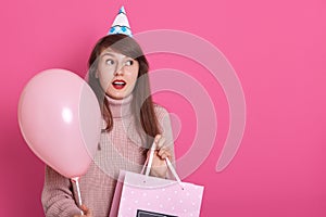 Happy brunette girl with pink balloon and bithday present in hands, keeps mouth opened, being in surprise, looks aside with