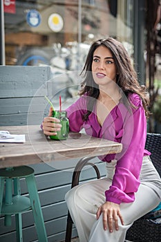 Happy Brunette Girl with pink blouse Sitting at the bar , Drinking a glass of lemonade While Smiling at the camera.