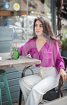 Happy Brunette Girl with pink blouse Sitting at the bar , Drinking a glass of lemonade While Smiling at the camera.