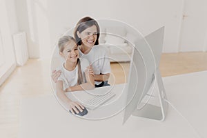 Happy brunette mother helps child do homework on computer, sit together at workplace, have cheerful expressions, focused into