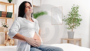 Happy brunette pregnant woman stroking belly enjoy peaceful tranquility relaxing on bed at home