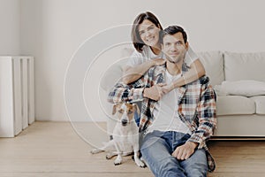 Happy brunette woman embraces husband with love, being in good mood, smiles positively. Husband, wife and dog pose together in